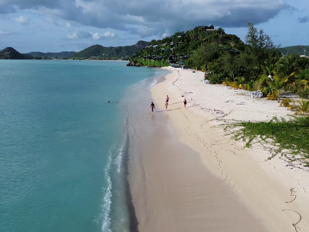 Aerial view of people walking the shoreline of a white-sand Antigua beach beside turquoise water