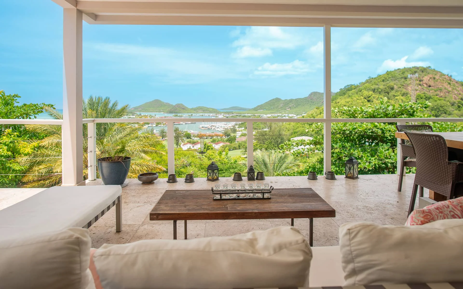 Covered villa terrace with a wooden table and lanterns, looking out over palms to Jolly Harbour and the green hills beyond